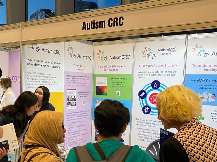 A group of people crowd around the Autism CRC booth, talking to Autism CRC staff and reading the booth infographics