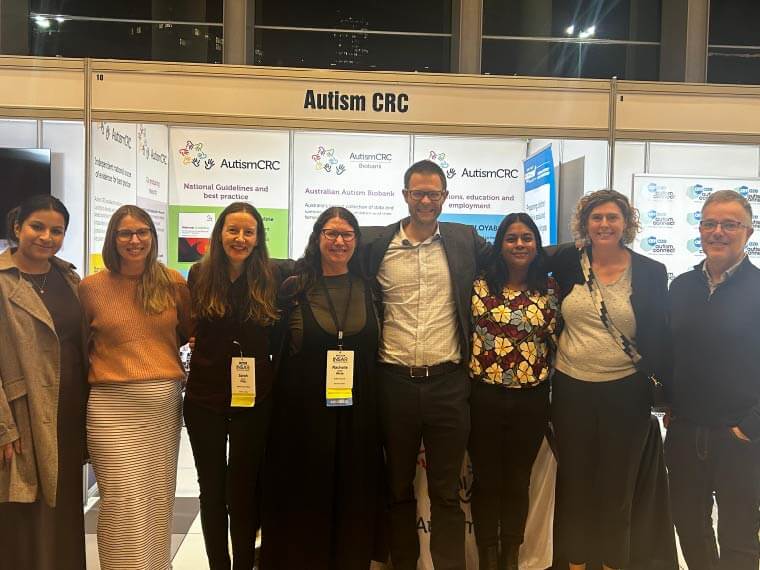 Eight people stand facing the camera in front of the Autism CRC booth